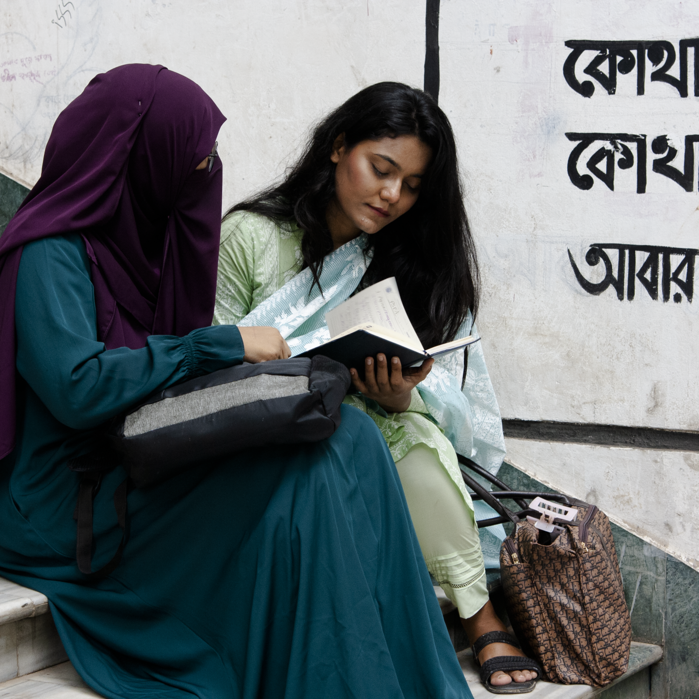 young women sit on steps one in a niquab one uncovered reading a book together. 