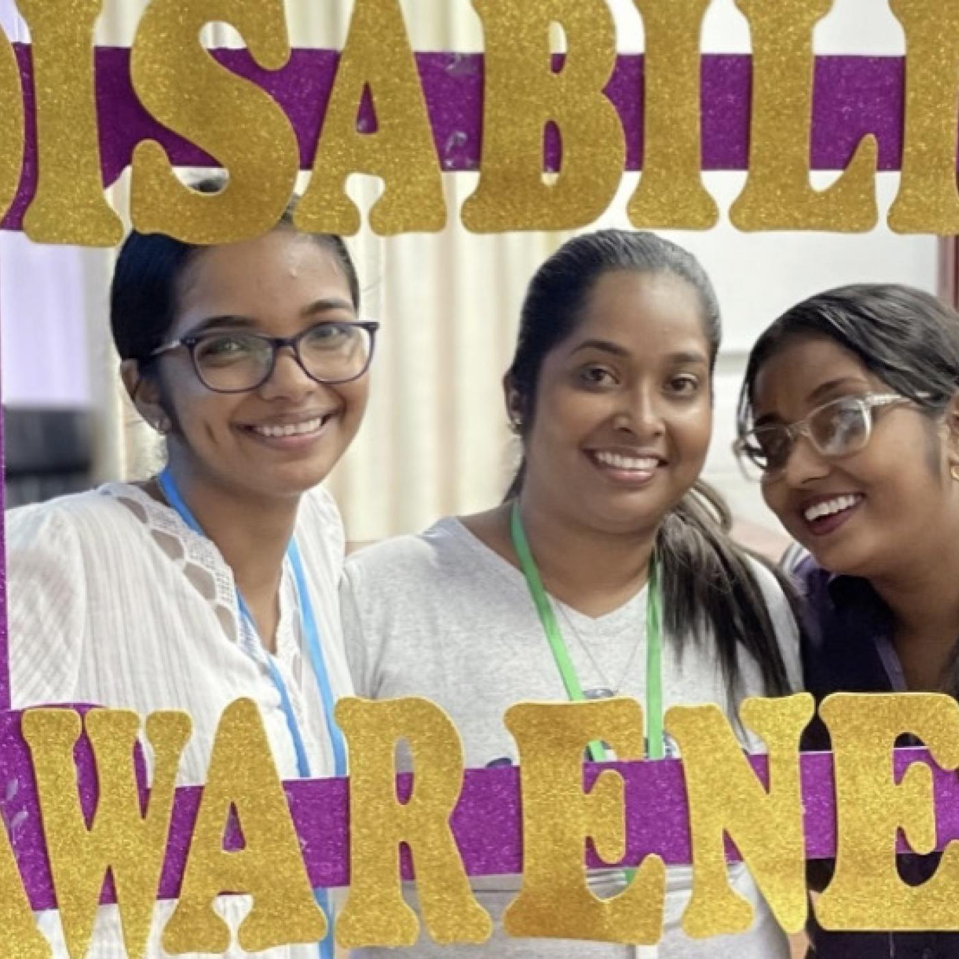Alt text: Three women from Guyana smile at the camera. They are holding up a purple photo frame that reads “Disability Awareness” in gold letters.
