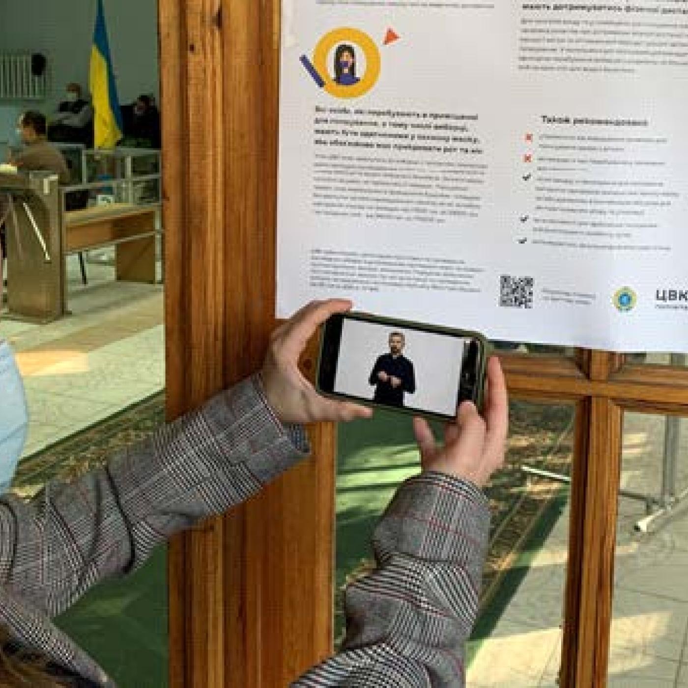 A woman holds up a smartphone to scan a QR code on a poster. Her smartphone plays a video of a person using sign language.